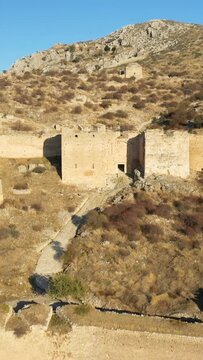 Stone fortress ruins stand on a rugged, sunlit hillside in Acrocorinth, Greece, with dry vegetation and rocky terrain under a clear blue sky.