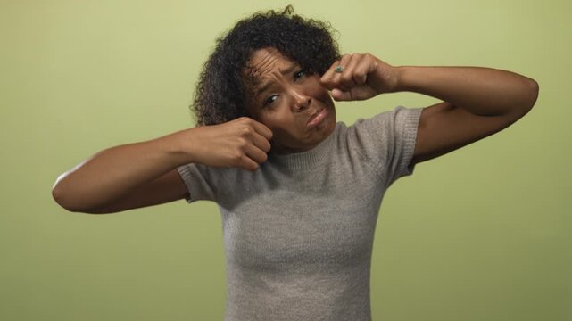 Young african american woman makes fists near eyes and pouts lips in studio portrait against pale green wall; sadness.