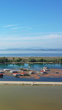 Aerial view of traditional stilt houses on calm blue water in Tourlida, Greece, with distant mountains and clear sky in the background.