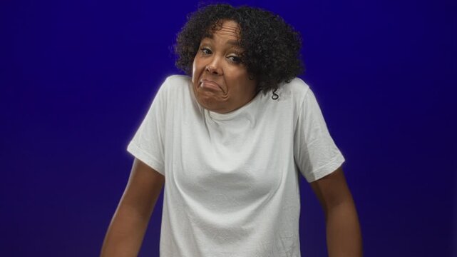 Young woman in white tshirt shrugs shoulders with puzzled expression in studio against blue backdrop; uncertainty.