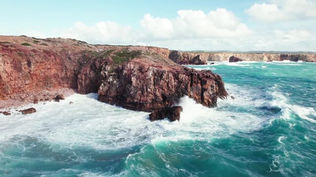 Powerful ocean waves crash against rugged reddish cliffs on windy day
