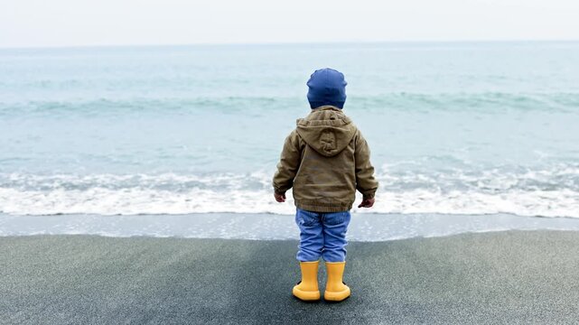 A boy in a jacket and rubber boots walks along the beach near the sea. A little 2 year old boy plays near the sea. The child breathes sea air.