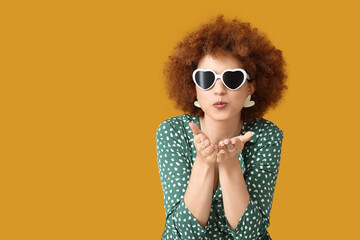 Redhead young woman with curly hair and sunglasses blowing kiss on orange background