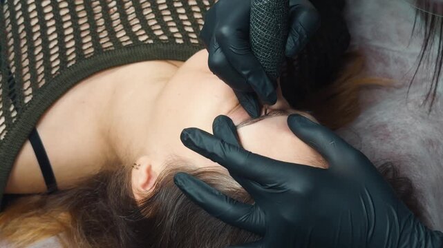 Woman receiving microblading procedure on eyebrows for permanent cosmetic makeup, enhancing facial features with semi permanent hairstrokes applied by a beauty professional wearing black gloves