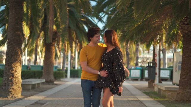 A couple strolls hand in hand along a sunny sidewalk lined with palm trees in the afternoon. They smile and share tender moments as they enjoy their time together in the city