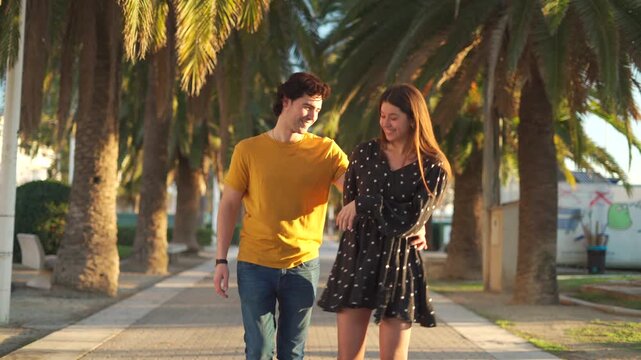 A man and woman walk together under palm trees in a park. They hold hands and share smiles, enjoying their time together in the afternoon sun