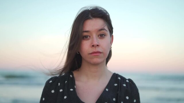 A young Hispanic woman stands by the ocean during sunset. She displays different feelings in four frames. Waves crash gently as the sky changes color. The mood shifts from serious to joyful