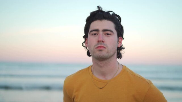 A young Hispanic man stands on the beach at sunset. He poses with different expressions. The ocean waves crash and the sun sets in the background. The sky shows colors of dusk