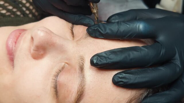 Cosmetologist's gloved hands applying microblading pigment, carefully drawing fine hair like strokes on a woman's eyebrows, enhancing beauty and facial features