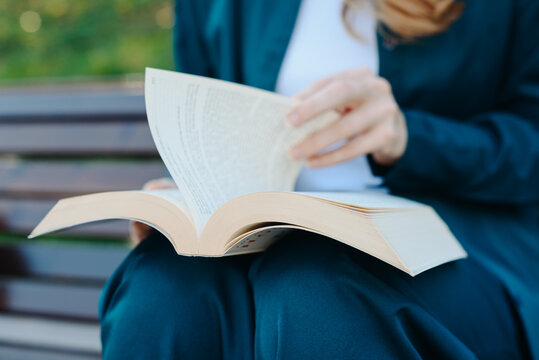 young woman holds thick book in hands, reading book, sitting on bench in park, close-up view, no face
