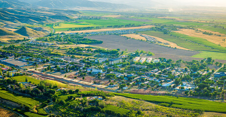Fototapeta premium Aerial view of a peaceful village nestled in a lush valley with vibrant agricultural fields and rolling mountains in Xinjiang, China.