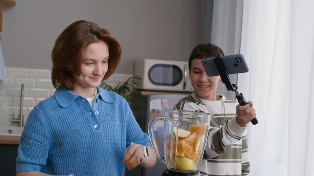 Eye level medium shot of two Caucasian teenagers preparing fruit smoothie in blender while recording video on smartphone with stabilizer in bright kitchen