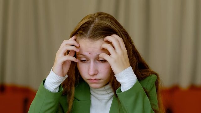 Stressed teenage girl in green blazer sitting at desk with notebook, holding her head in hands and bending over table, concept exam stress and school burnout in modern students.