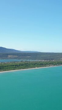 Aerial shot of turquoise sea, sandy beach, and lush greenery at Isola Varano Nature Reserve in Italy. Clear blue sky and distant hills create a serene natural landscape.