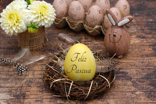 Un huevo amarillo con la inscripci&oacute;n en espa&ntilde;ol "Feliz Pascua" en un nido de ramitas y plumas, junto a &eacute;l un conejito de Pascua de madera marr&oacute;n, una huevera con huevos de Pascua y un jarr&oacute;n con flore