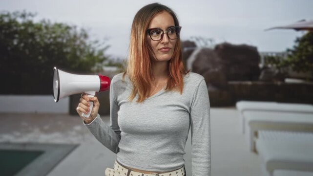 Young redhead woman holds megaphone near poolside lounge chairs and rock fountain at hotel building, wearing glasses and grey sweater; confidence announcement.