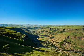 Obraz premium Wide green valley under clear blue sky, rolling hills, bright sunlight, distant ridgelines, tranquil open landscape ideal