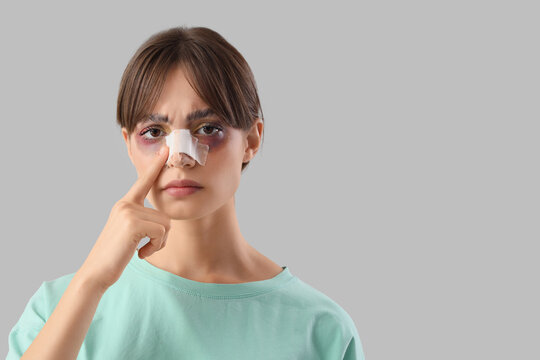 Beautiful young worried woman touching her bandaged nose on grey background. Plastic surgery concept