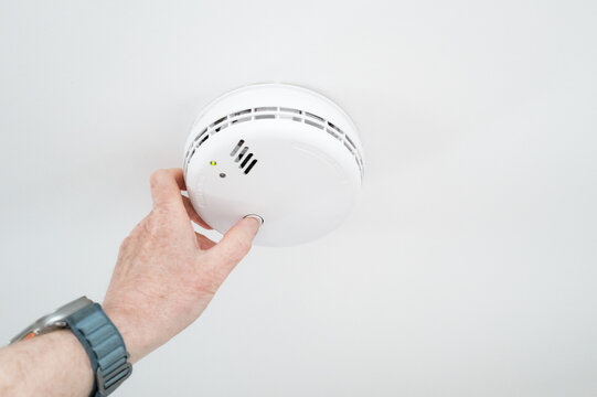 Homeowner seen conducting a weekly siren test on a ceiling mounted smoke and carbon monoxide detector located in a hallway. The detector is mains powered and the constant green LED light can be seen.
