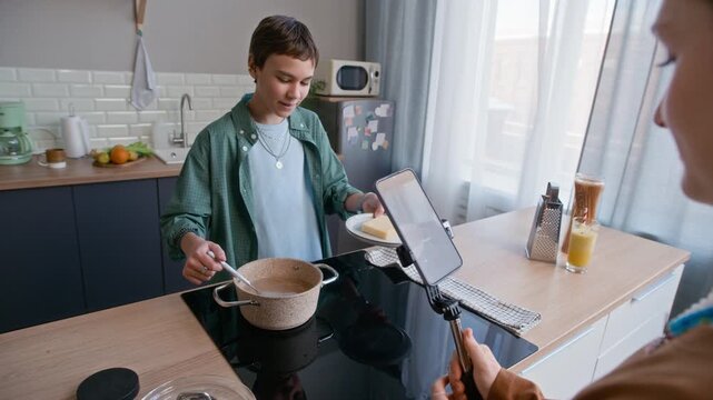 Over shoulder of two Caucasian teenagers using smartphone on stabilizer to film cooking video, boy stirring spaghetti in pot while girl recording in bright modern kitchen