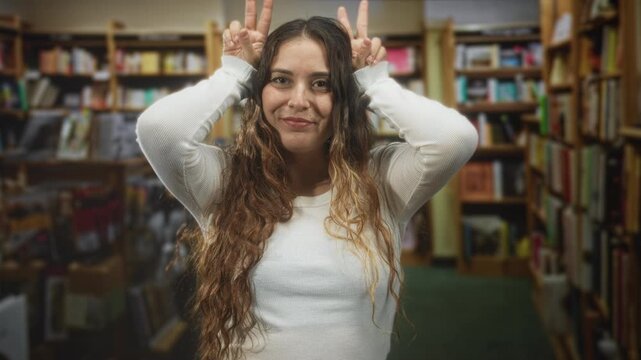 Woman sticking out tongue and making bunny ears with hands amid crowded bookshelves in building; playful mischief.