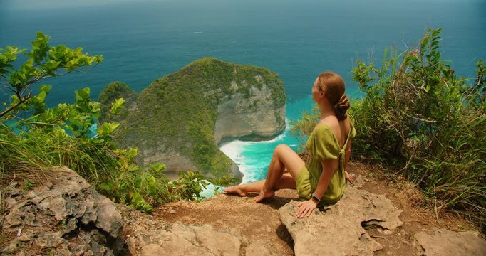 Tourist woman sits on rock watching ocean waves at Kelingking Beach coastal landmark in Nusa Penida Bali
