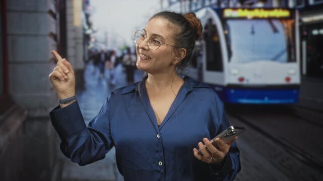 Woman brunette points finger while holding smartphone on street by tram and tracks, smiling with glasses and blue shirt; curiosity discovery.