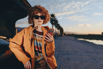 A woman with curly hair wearing sunglasses and a rainbow sweater stands next to an open car trunk while using a smartphone outdoors during sunset. Lifestyle concept. © SHOTPRIME STUDIO