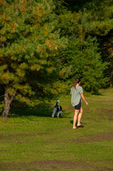 Fototapeta premium A woman walks a leashed dog on a grass field beside a forest during a sunny day in the summer.