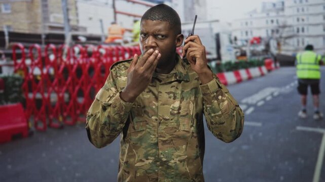 Young black soldier man covering mouth with hand while speaking into radio on urban street during roadworks; concern duty.