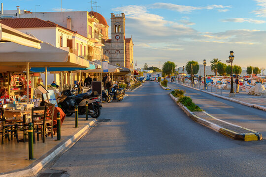 Shops and sidewalk cafes along the harbor waterfront of the town of Aegina, Greece, with Saint Nicholas church in view on the Saronic Greek island of Aegina.