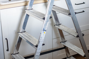 Aluminum step ladder positioned near white kitchen cabinets with black handles and a sink, showcasing a home improvement setting for electrical or maintenance tasks © Irina Flamingo