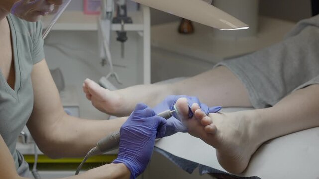 Closeup salon electric pedicure shaping toenails with rotary drill and blue gloves, calm client relaxation