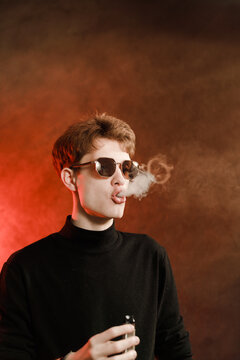 Young man in black turtleneck and sunglasses exhaling smoke vape, studio portrait on dark background.
