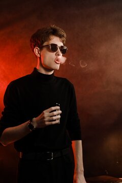 Young stylish man in black turtleneck and sunglasses exhaling smoke rings from vape, studio portrait on dark background with red light.