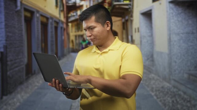 Man holding laptop typing with right hand in a narrow cobblestone street while checking the screen in a yellow polo; concentration remote work.