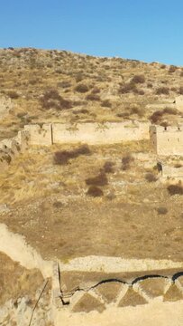 Historic stone walls and ruins stretch across a dry, rocky hillside under a clear blue sky at Acrocorinth, Greece.