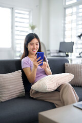 Obraz premium Asian adult woman uses mobile phone online communication social media browsing at modern cozy home living room relaxing comfortable environment happy digital lifestyle technology connection