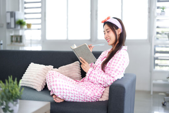 Asian adult woman writing journal on living room sofa wearing pajamas relaxing planning future thoughts enjoying peaceful home comfort