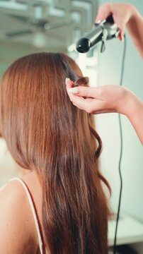 Close-up from behind of hairdresser hand use curling iron to create curls on client long, healthy brown hair in salon. Professional salon styling for special event, beauty routine, precision and care