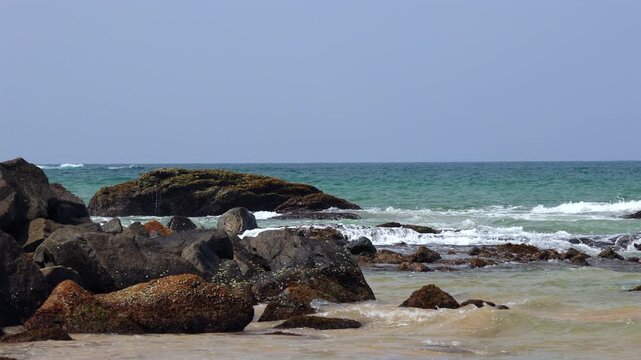 Ocean Waves Rushing and Splashing Against Moss-Covered Rocks on a Sandy Shoreline with a Clear Blue Sky Horizon View. Sri Lanka island in Asia.
