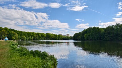 Willows grow on the grassy shore of the lake. There is a park on the shore, and behind it there are buildings. There are ripples and reflections of the sky on the water, and buoys installed. Sunny © Balser