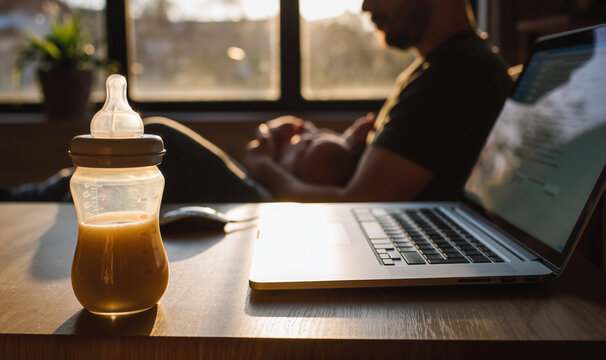 Warm baby bottle with formula milk propped next to a working laptop, and a young father caring and feeding his infant in background. Modern fatherhood concept as balance of parenting and remote work