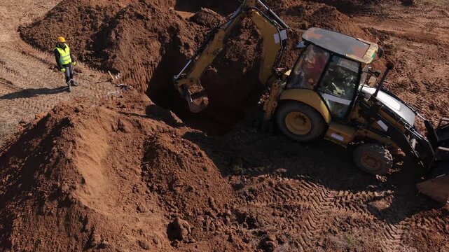 Aerial View of a Yellow Backhoe Excavator Digging a Foundation Pit Supervised by a Construction Worker