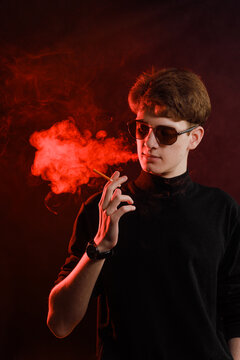 Stylish young man in black turtleneck and sunglasses smoking a cigarette, studio portrait with red neon lighting and dramatic smoke.