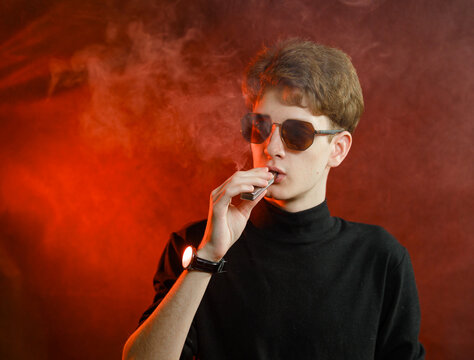 Young man smoke from vape, studio portrait on dark background with night red light.