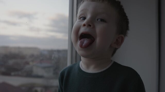 Funny child making faces, sticking out tongue and playing near the window at home