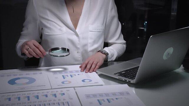 Person analyzing business data with magnifying glass, scrutinizing financial charts for auditing and research. Audit in business and overworking concepts