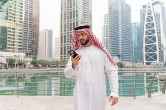 A confident Arabic businessman checks his smartphone in urban area during daytime, representing modern business in the region.