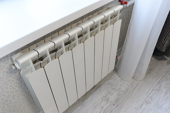 A white bi-metal radiator with 10 sections is mounted under a window, featuring a red valve, adjacent to light curtains and a leopard-print wallpaper background.